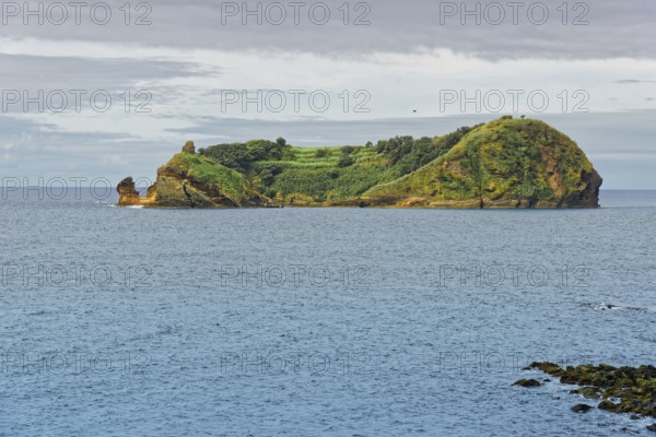 Green, rocky island and protected area Ilhéu de Vila Franca do Campoin a calm sea under a cloudy