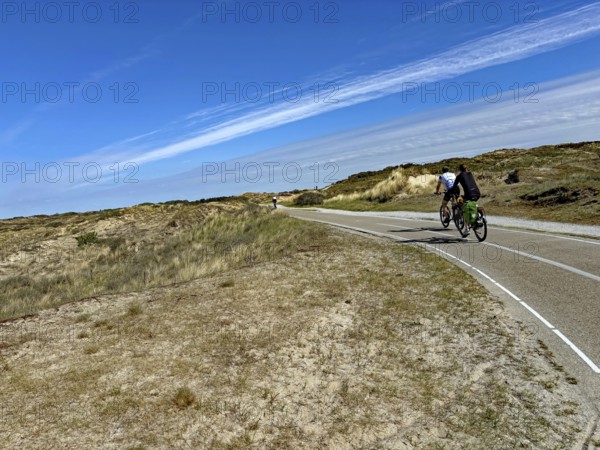 Cyclist on a wide cycle path through a dune landscape under a blue sky with clouds, north sea Cyle