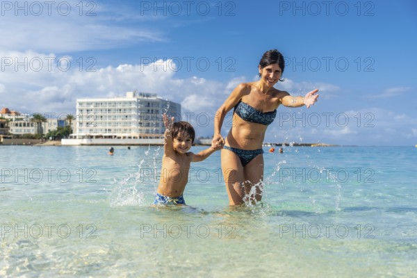 Mother and son holding hands and splashing in the crystal clear water of playa de s'estany d'en mas