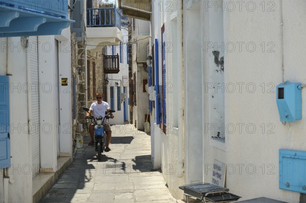 A narrow white alley with blue shutters. A man rides a motorbike through the sun, Mandraki,