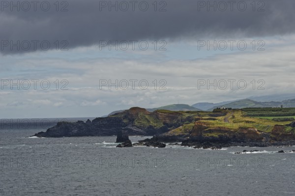 A rocky coastline with green hills in the background under a cloudy sky, Fenais da Luz, Sao Miguel