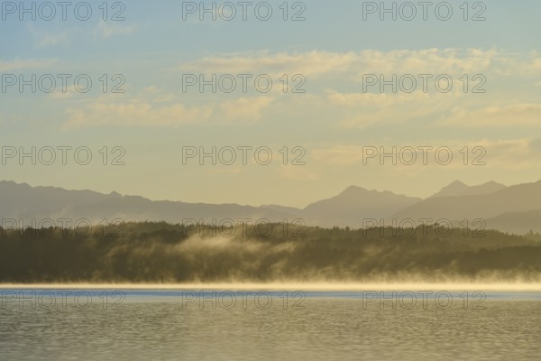 Fog over a calm lake under a clear sky with mountains in the background, Lake Mahinapua, Ruatapu,