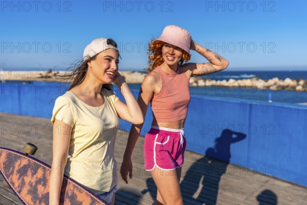 Two young women walk along a boardwalk by the sea, enjoying a sunny day. One carries a skateboard,