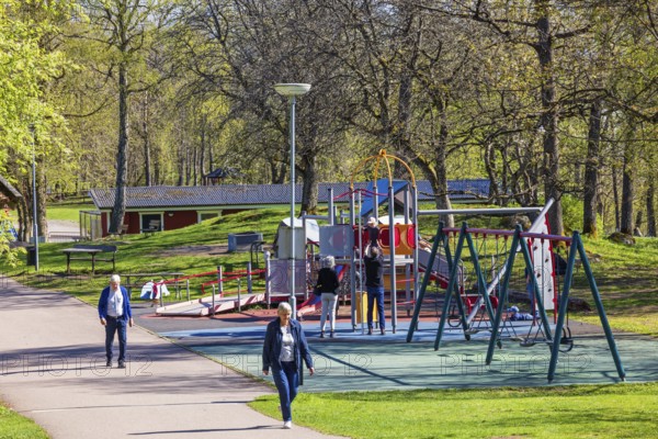 Playground in a public park with children playing and people walking on a footpath at springtime,