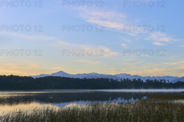 A calm, misty lake in morning sunlight with mountains in the background, Lake Mahinapua, Ruatapu,