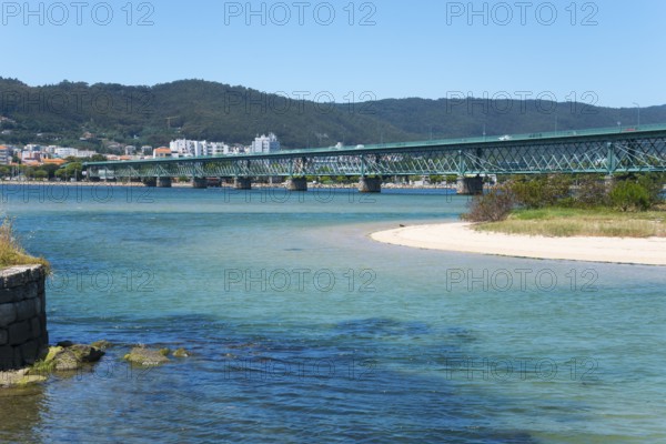 A long bridge crosses a shimmering blue river, surrounded by hills and a town in the background,