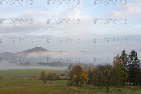Fog in the valley, surrounded by trees and a small house, with a range of hills in the background,