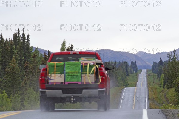 Red lorry with bulky goods drives up and down a straight road lined with forests and mountains,