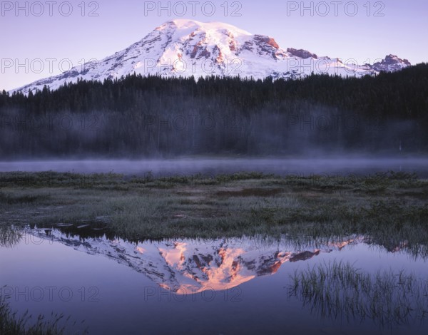 Snow capped mountain reflecting in a calm lake at dusk, surrounded by mist and lush forest. The sun