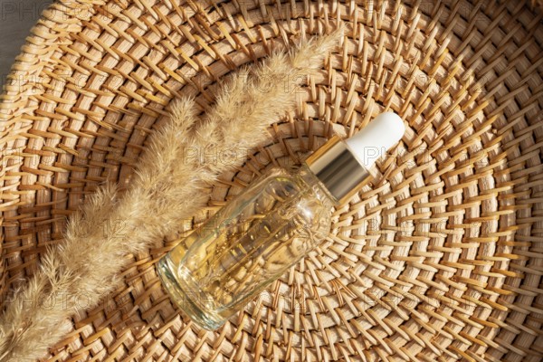 A glass cosmetic with golden essential oil rests on a woven basket alongside dried grass. The
