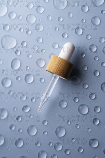 A glass pipette bottle lies on a textured blue surface Water droplets surround the bottle,