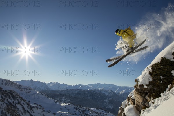 A skier in yellow gear performs a thrilling jump off a cliff in a sunny, snow-covered alpine
