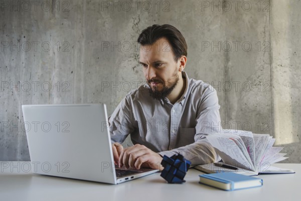 A man in a shirt works intently on a laptop at office. Beside him are a book and a Rubik cube,