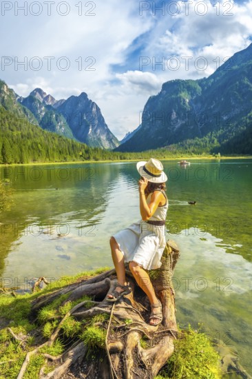 Young woman wearing a straw hat sitting on a tree stump by lake dobbiaco, enjoying the breathtaking