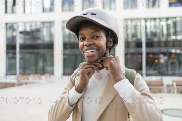 A black woman smiles while adjusting her helmet in an urban setting She is dressed modernly,