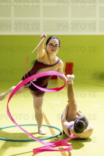 Two disabled rhythmic gymnasts perform with a pink ribbon and hoop in a vibrant indoor space. One