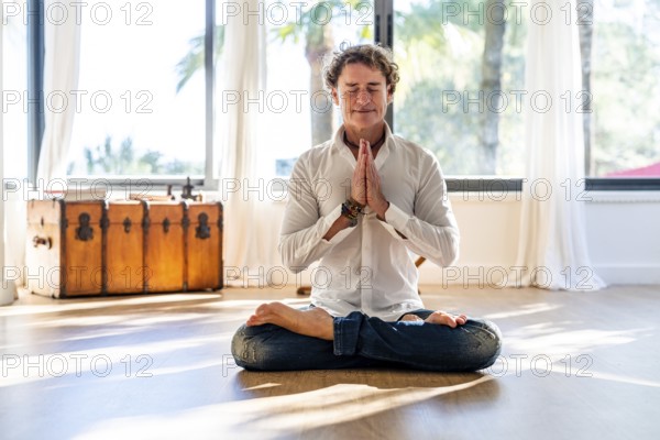 A calm man with curly hair meditates with eyes closed, seated in a lotus pose, hands in prayer