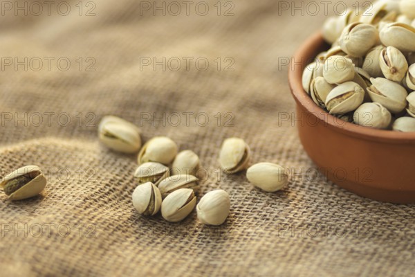 Close up of a clay bowl filled with pistachios on a burlap surface. Several pistachios are