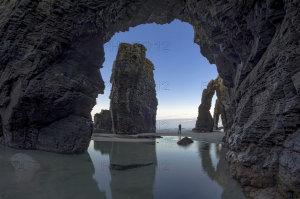 A person stands between towering sea arches, reflected in calm tidal pools, under a serene sky. The