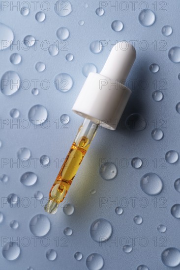 A close-up of a pipette filled with vitamin C serum on a blue surface decorated with water droplets