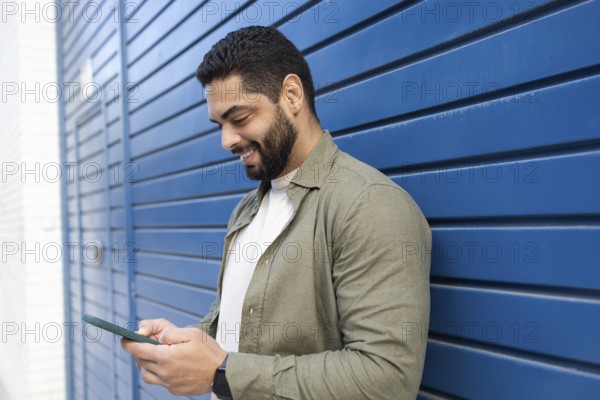 A man stands against a blue wall, smiling as he looks at his smartphone in the city. He wears a
