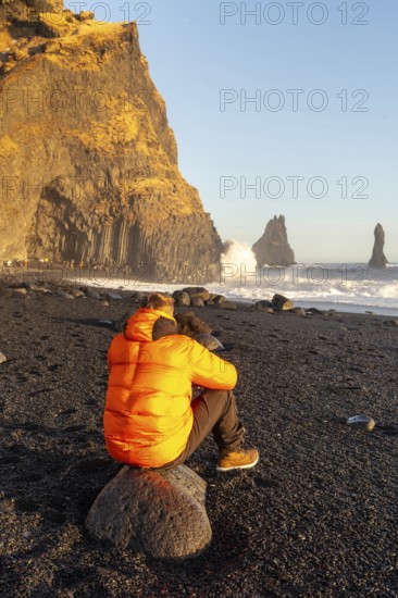 A man in an orange jacket sits on a rock on a beach. The sky is blue and the sun is shining. The