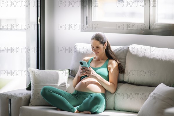 A pregnant woman in a green yoga outfit relaxes on a comfortable couch, focused on her smartphone.