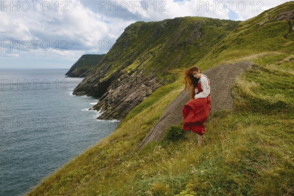 A young woman wearing a flowing red dress and white blouse explores the rugged, green hillside