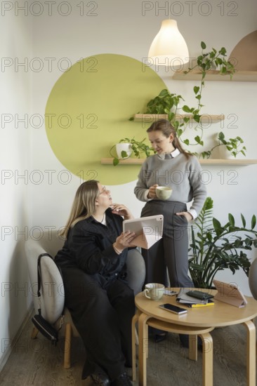 Two women engage in conversation in a cozy cafe setting. One sits with a tablet while the other