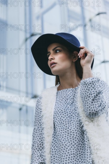 Elegant woman wearing a blue hat and fur vest, looking poised in an urban setting with glass