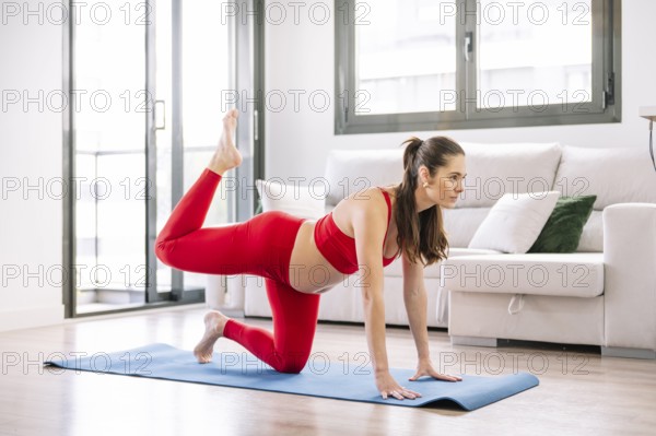 A pregnant woman in a red sports outfit practicing yoga on a blue mat indoors. She performs a pose
