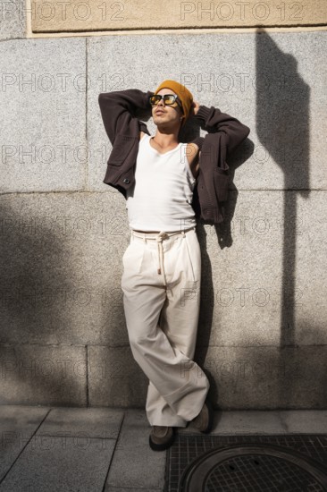 Stylish Colombian gay man wearing sunglasses and a beanie, leans against a city wall. His casual