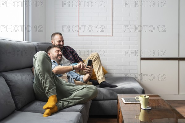 A happy gay couple relaxes on a cozy couch, smiling as they look at a smartphone. The setting is a