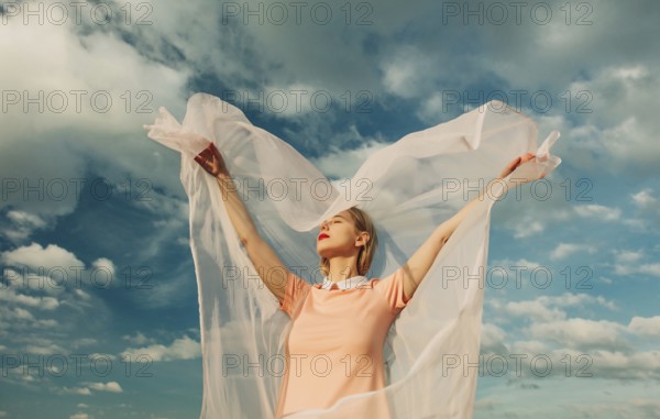 A woman with outstretched arms embraces the breeze, her sheer fabric flowing gracefully. A serene