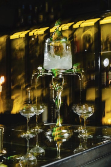 A vintage absinthe fountain pours into glasses on a dark bar counter. The ambiance is enhanced by a
