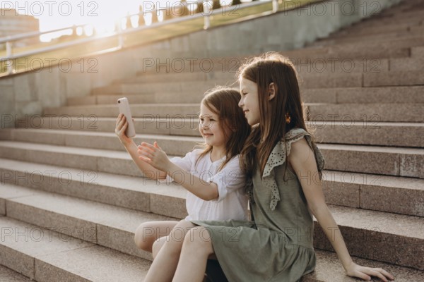 Two young girls sit on sunlit steps in summer, happily taking a selfie. They are enjoying a sunny