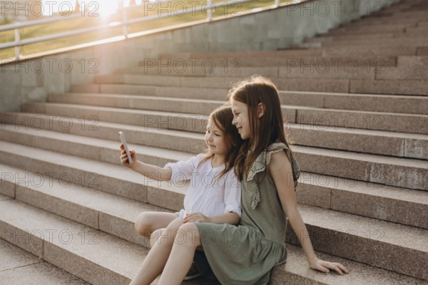 Two young girls sit together on sunlit stone stairs, smiling as they take a selfie. The warm glow