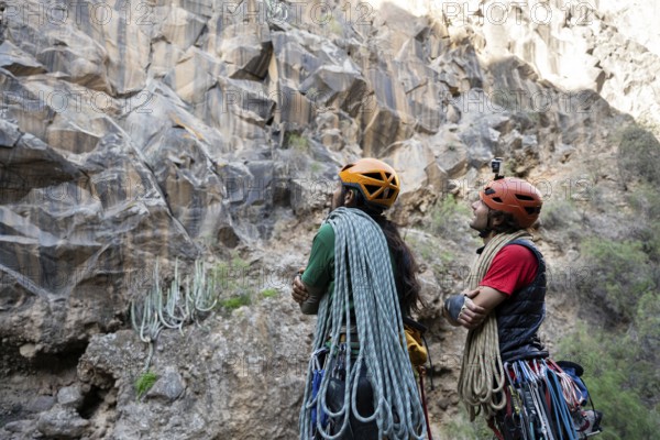 Two climbers equipped with helmets and ropes study a rugged cliff, preparing for a traditional