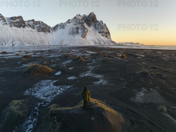 A man stands on a textured, frost-covered dune, gazing at the towering, snow-draped mountains of
