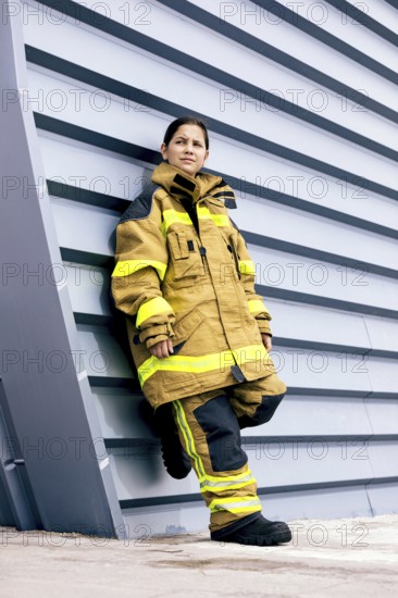 A young girl stands confidently against a modern wall while wearing a firefighter suit, showcasing