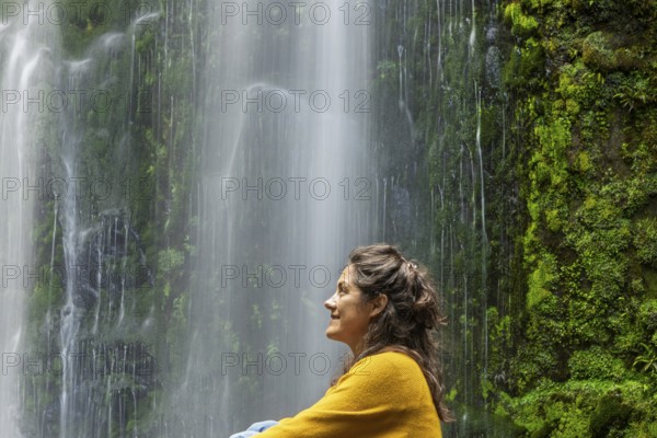 A woman in a yellow sweater enjoys a tranquil moment beside a lush green waterfall, surrounded by