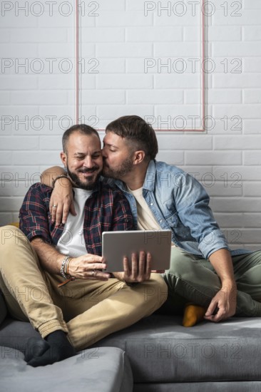 A loving gay couple sits on the couch, sharing a sweet moment while using a tablet. They embrace