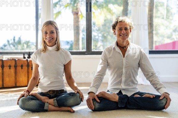 Male and female couple sits cross-legged on the floor in a sunlit room, meditating looking at