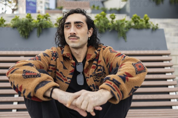 A man with curly hair and a mustache sits on an outdoor bench wearing a patterned jacket and