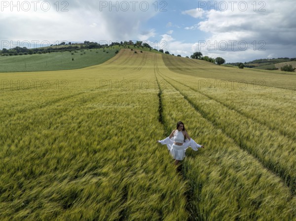 A young woman strolls through lush green fields under a wide blue sky in rural Italy, embracing the