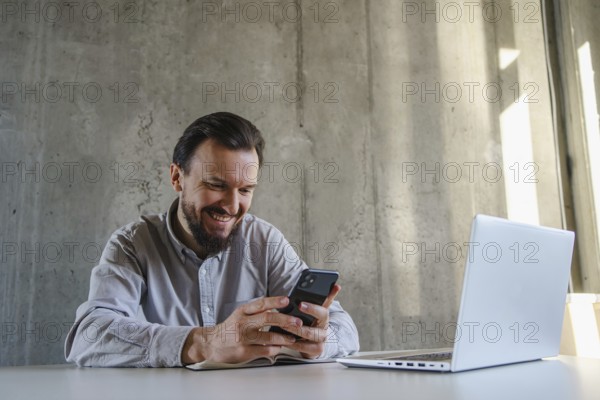 A man in a casual shirt sits at a desk, smiling while using his smartphone. Nearby, a laptop is