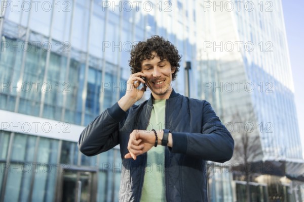 Smiling man in casual attire checks the time on his smartwatch while talking on a smartphone.