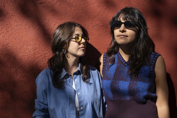 A multiethnic lesbian couple shares a moment outdoors, standing against a sunlit red wall. Both
