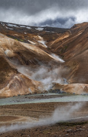 Steaming stream between colourful rhyolite mountains with snowfields, Hveradalir geothermal area,