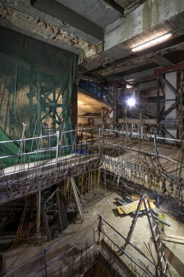 An interior construction site with exposed beams, scaffolding, and safety nets. Illuminated by